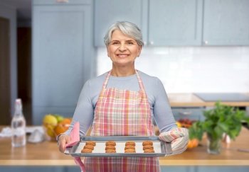 cooking, culinary and old people concept - smiling senior woman in apron with cookies on baking pan over home kitchen background. senior woman in apron with cookies on baking pan