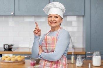 cooking, culinary and old people concept - portrait of smiling senior woman or chef in toque in apron pointing finger up over home kitchen background. smiling senior woman or chef pointing finger up