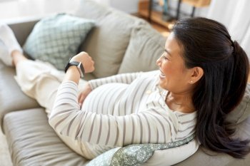 pregnancy, rest, people and expectation concept - happy smiling pregnant asian woman with smart watch sitting on sofa at home. happy pregnant woman with smart watch at home