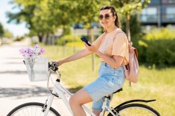 people, leisure and lifestyle - happy young woman with smartphone, backpack and flowers in basket of bicycle on city street. woman with smartphone on bicycle in city