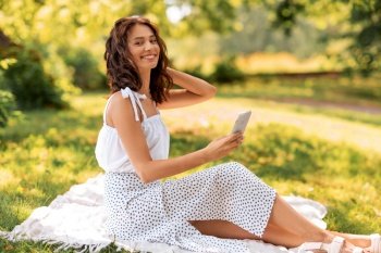 leisure and people concept - happy smiling young woman with smartphone sitting on picnic blanket at summer park. happy smiling woman with smartphone summer at park