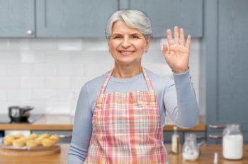 cooking, culinary and old people concept - portrait of smiling senior woman in apron waving hand over home kitchen background. smiling senior woman in apron waving hand