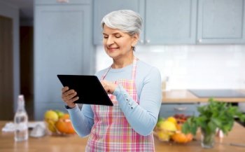 cooking, culinary and old people concept - portrait of smiling senior woman in apron with tablet pc compute over home kitchen background. smiling senior woman in apron with tablet computer