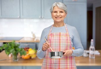 food cooking, culinary and old people concept - portrait of smiling senior woman in kitchen apron with pot and spoon over home kitchen background. senior woman in apron with pot cooking food
