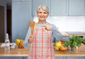 food cooking, culinary and old people concept - portrait of smiling senior woman in apron with wooden spoons over home kitchen background. smiling senior woman in apron with wooden spoons
