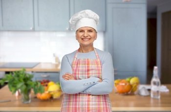 cooking, culinary and old people concept - portrait of smiling senior woman or chef in toque in apron with crossed arms over home kitchen background. smiling senior woman or chef in toque at kitchen