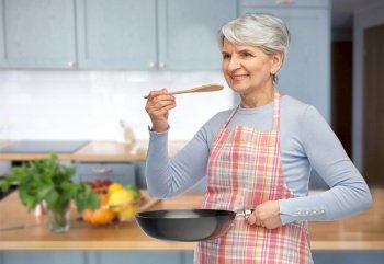 cooking, culinary and old people concept - portrait of smiling senior woman in apron with frying pan and spoon tasting food over kitchen background. smiling senior woman in apron with frying pan