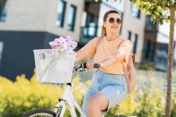 people, leisure and lifestyle - happy smiling young woman with earphones, backpack and flowers in basket riding bicycle on city street. happy woman with earphones riding bicycle in city