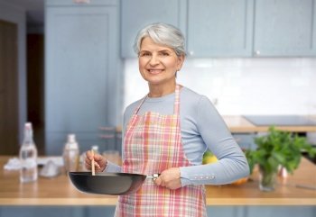 food cooking, culinary and old people concept - portrait of smiling senior woman in apron with frying pan over kitchen background. smiling senior woman in apron with frying pan