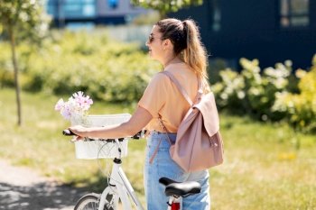 people, leisure and lifestyle - happy young woman with flowers in basket of bicycle and backpack on city street. woman with flowers in bicycle basket in city