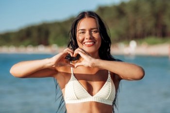 people, summer and swimwear concept - happy smiling young woman in bikini swimsuit showing hand heart gesture on beach. smiling young woman in showing hand heart on beach