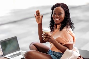 technology, education and people concept - happy smiling african american student girl with takeaway coffee cup and laptop computer waving hand in city. african student girl with coffee cup and laptop