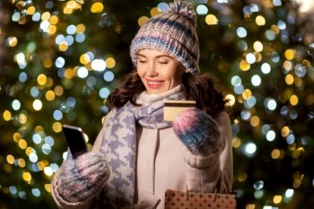 winter holidays, season sale and people concept - happy smiling young woman with christmas gifts in shopping bags, credit card and smartphone over festive lights. woman with phone, credit card and christmas gifts