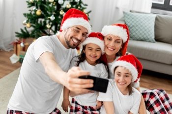 family, winter holidays and christmas concept - happy mother, father and two daughters in santa hats at home taking selfie with smartphone. happy family taking selfie on christmas at home