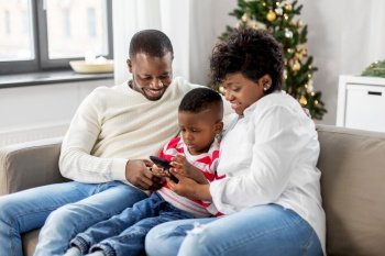 family, winter holidays and people concept - happy african american mother, father and little son with smartphone at home on christmas. african family with phone on christmas at home