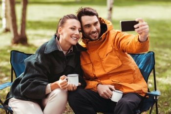 camping, tourism and travel concept - happy couple with smartphone drinking tea and taking selfie at tent camp. couple drinking tea and taking selfie at tent camp