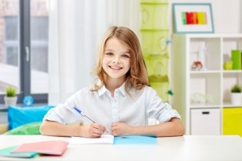 education, elementary school, learning and people concept - happy smiling girl with notebook and pen sitting at table over home background. happy smiling school girl with notebook and pen