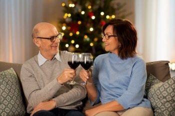 winter holidays and people concept - happy smiling senior couple toasting glasses of red wine at home in evening over christmas tree lights on background. happy senior couple drinking red wine on christmas