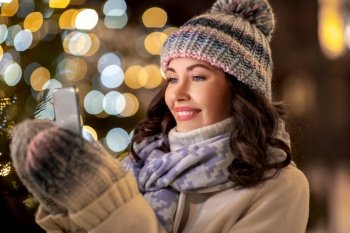 holidays, communication and people concept - portrait of beautiful happy smiling young woman using smartphone or taking selfie over christmas lights in winter city. happy woman with smartphone in christmas city