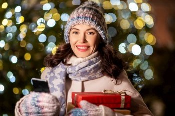 winter holidays, celebration and people concept - happy woman with christmas gifts and smartphone over festive lights. happy woman with christmas gifts and smartphone