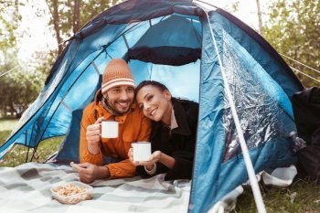 camping, tourism and travel concept - happy couple lying inside tent and drinking tea at campsite. couple drinking tea lying inside tent at campsite