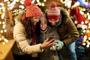 family, winter holidays and technology concept - happy mother, father and little daughter with smartphone at christmas market in evening. happy family with smartphone at christmas market