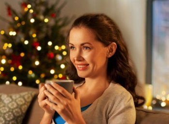 people, holidays and leisure concept - happy woman with mug drinking tea or coffee at home in evening over christmas tree lights on background. happy woman drinking tea or coffee on christmas