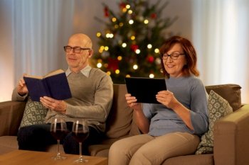 leisure and winter holidays concept - happy senior couple reading book and using tablet pc computer at home in evening over christmas tree lights on background. senior couple with book and tablet pc on christmas