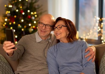 technology, winter holidays and people concept - happy senior couple taking selfie with smartphone at home in evening over christmas tree lights on background. old couple taking selfie with phone on christmas