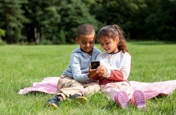 childhood, leisure and technology concept - happy children with smartphone sitting on blanket outdoors. kids with smartphone sitting on blanket outdoors