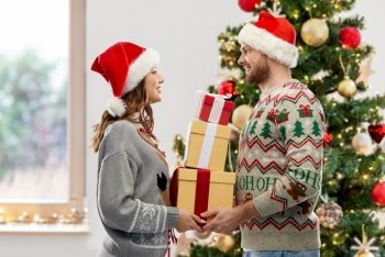 people and holidays concept - happy couple in santa hats with gifts at ugly sweater party over christmas tree on background. happy couple in christmas sweaters with gifts