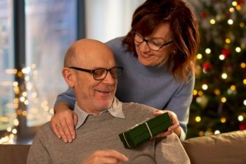 winter holidays and people concept - happy senior couple with gift box at home over christmas tree lights on background. happy senior couple with christmas gift at home