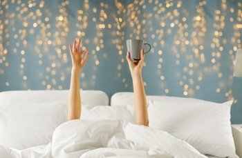 morning, comfort and people concept - young woman with cup of coffee lying in bed and showing ok hand sign over festive lights on background. woman with cup of coffee lying in bed showing ok
