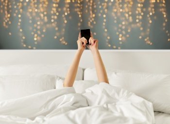 technology, comfort and morning concept - hands of young woman lying in bed with smartphone over festive lights on background. hands of woman lying in bed with smartphone