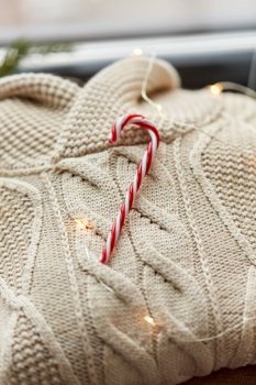 christmas, clothes and objects concept - close up of warm wool braided sweater, candy cane and electric garland string at home. close up of sweater, candy cane, christmas garland