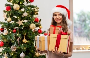 winter holidays and people concept - happy smiling teenage girl in santa helper hat holding gift box over christmas tree on background. teenage girl in santa hat with christmas gift