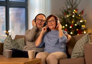 winter holidays, leisure and people concept - happy senior couple with headphones listening to music at home in evening over christmas tree lights on background. senior couple listening to music on christmas