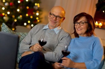 winter holidays, leisure and people concept - happy smiling senior couple with glasses of red wine watching tv at home in evening over christmas tree lights on background. couple drinks wine and watches tv on christmas