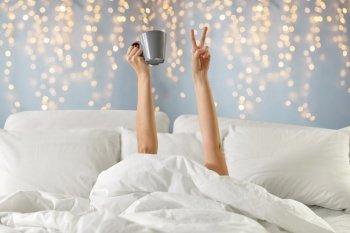 morning, comfort and people concept - young woman with cup of coffee lying in bed and showing peace hand sign over festive lights on background. woman with coffee lying in bed showing peace