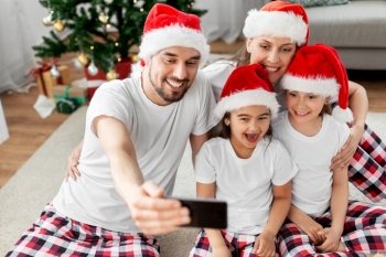 family, winter holidays and christmas concept - happy mother, father and two daughters in santa hats at home taking selfie with smartphone. happy family taking selfie on christmas at home