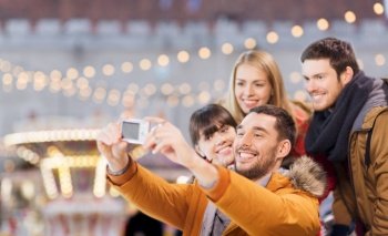 winter holiday, technology and leisure concept - happy friends taking selfie with digital camera over christmas market or amusement park background. friends taking selfie with camera on christmas