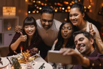 holidays, party and celebration concept - multiethnic group of happy friends having christmas dinner at home and taking selfie with smartphone. friends taking selfie on christmas dinner at home