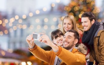 winter holidays, technology and leisure concept - happy friends taking selfie with digital camera over christmas market background. happy friends with camera taking christmas selfie