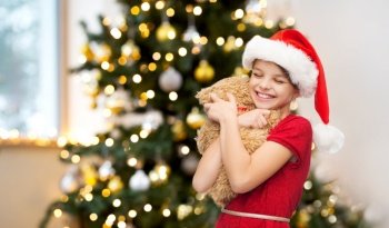 winter holidays, childhood and happiness concept - smiling girl in santa helper hat with teddy bear over christmas tree background. smiling girl in santa helper hat with teddy bear