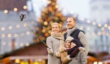 winter holidays, technology and people concept - happy family taking picture with smartphone and selfie stick over christmas market background. happy family taking selfie at christmas market