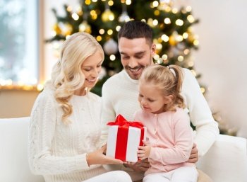 family, winter holidays and people concept - happy mother, father and little daughter with gift box sitting on sofa at home over christmas tree background. happy family at home with christmas gift box