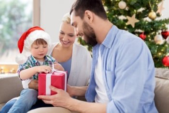 family, winter holidays and people concept - happy mother, father and little son with gift box sitting on sofa at home over christmas tree background. happy family at home with christmas gift box
