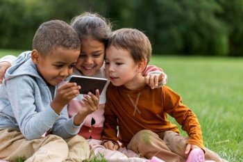 childhood, leisure and technology concept - happy children with smartphone sitting on blanket at park. kids with smartphone sitting on blanket at park