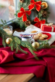christmas and winter holidays concept - camp cup of coffee with candy cane, oatmeal cookies on red ceramic plate, gift and decorations on window sill at home. cup of coffee, cookies and christmas decor at home