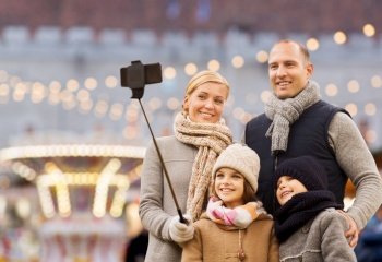 winter holidays, technology and people concept - happy family taking picture with smartphone and selfie stick over christmas market or amusement park background. happy family taking selfie at christmas market